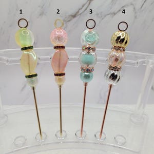 May include: Four decorative metal pins labelled 1 through 4, each featuring a unique arrangement of colourful beads and rhinestones. The pins have a thin, pointed metal shaft and a circular loop at the top, displayed upright in a clear acrylic stand.
