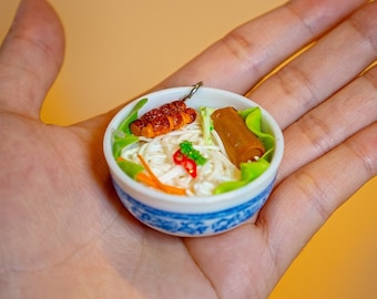 May include: A miniature bowl of white noodles with a small piece of grilled meat, a spring roll, and green vegetables. The bowl is decorated with blue and white floral patterns.