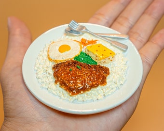 May include: A miniature white plate with a small portion of rice, a fried egg, a piece of meat, and a small piece of cheese. The plate is held in a person's hand.