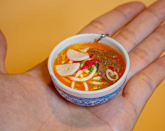 May include: A small, white bowl with blue trim filled with a miniature version of a noodle soup. The soup contains noodles, meat, and vegetables. The bowl is being held in a person's hand.