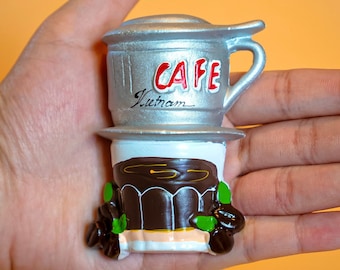 May include: A silver and brown ceramic figurine of a Vietnamese coffee maker. The coffee maker has a silver top with the word "CAFE" and "Vietnam" written on it. The bottom of the coffee maker is brown and has a white base.