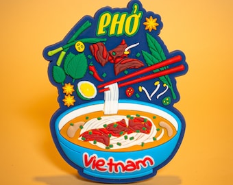 May include: A blue and yellow rubber magnet with a cartoon illustration of a bowl of Pho, a Vietnamese noodle soup. The magnet says "Pho" and "Vietnam" in red letters.