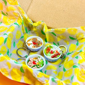 May include: Three miniature food bowl keychains with detailed food replicas, including noodles, vegetables, and meat. The keychains are in a cardboard box lined with lemon-patterned tissue paper. The background is a bright orange.