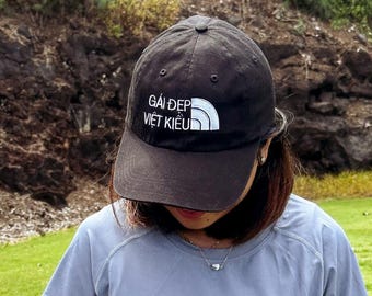 May include: A black baseball cap with the white text GÁI ĐẸP VIỆT KIỀU, a light blue short-sleeved athletic shirt, and a white athletic skirt. The outfit includes a white golf glove on one hand and a pink golf club grip is visible in the foreground.