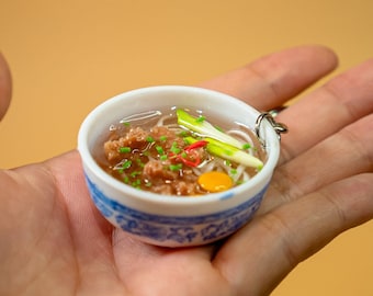 May include: Miniature ceramic bowl of soup with noodles, meat, and vegetables. The bowl has a blue and white floral design. The soup contains green onions, red pepper, and a yellow yolk. A silver ring is attached to the bowl.