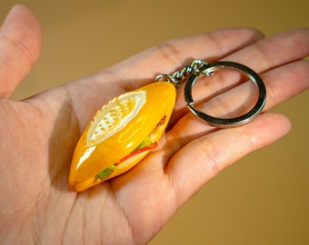 May include: A yellow and green keychain shaped like a baguette sandwich. The keychain has a silver metal ring.