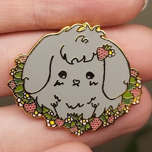 May include: A grey enamel pin of a fluffy bunny wearing a wreath of pink and white flowers and strawberries. The bunny has a heart-shaped nose and closed eyes.