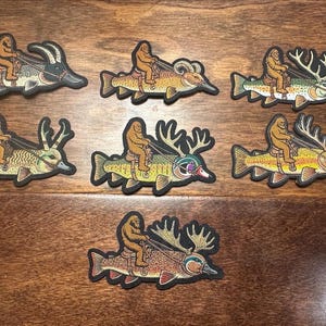May include: Seven embroidered patches featuring a brown Bigfoot figure riding various fish, including pike and trout, with some fish sporting antlers or horns. Each patch has a black border and depicts a creature saddled and bridled on a fish.