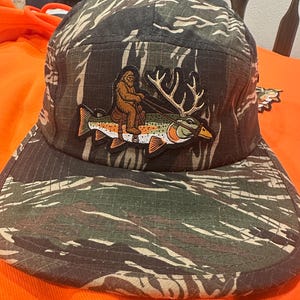 May include: A five-panel cap featuring a tiger stripe camouflage pattern in shades of green, brown, and black. Centered on the front is an embroidered patch depicting a brown Bigfoot figure riding a large fish with prominent deer antlers.