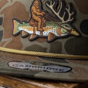 May include: A camouflage baseball cap featuring an embroidered patch of a brown Bigfoot figure riding a large fish with antlers. The cap includes a gold braided cord across the base of the crown and a sticker on the brim with the text CAMBRIDGE.