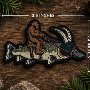 May include: An embroidered patch featuring a brown Bigfoot figure riding a large fish with goat horns and a bridle. The patch measures 3.5 inches in width and 2.4 inches in height, with a black border and detailed stitching in green, tan, and white.