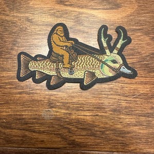 May include: An embroidered patch featuring a tan Bigfoot figure riding a large fish with antlers. The fish has a patterned body, a saddle, and a bridle. The design is outlined in black thread, showcasing a mythical creature and a fish hybrid.