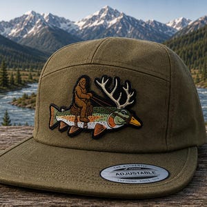 May include: An olive green five-panel cap featuring a central embroidered patch of a brown Bigfoot figure riding a large rainbow trout with deer antlers. The cap has a flat brim and a circular sticker on the brim that reads The Authentic Adjustable.