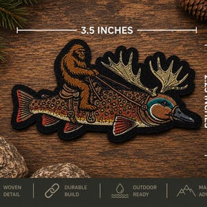 May include: A woven patch featuring a brown Bigfoot figure riding a large fish with moose antlers. The patch measures 3.5 inches in width and 2.25 inches in height. The design includes a saddle and bridle on the fish, with detailed embroidery.