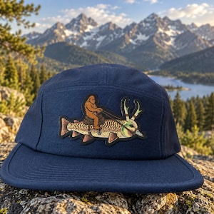 May include: A navy blue five-panel cap featuring a central patch illustration of a brown Bigfoot creature riding a large fish with antlers. The fish has a bridle and reins attached, and the cap has a flat brim and a structured crown design.