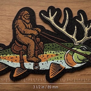 Op de afbeelding: Een geborduurde patch met een bruine Bigfoot-figuur die op een groen-oranje vis met een groot gewei rijdt. De patch is 52,3 mm hoog en 89 mm breed, zoals aangegeven door de zichtbare maataanduidingen op de afbeelding.