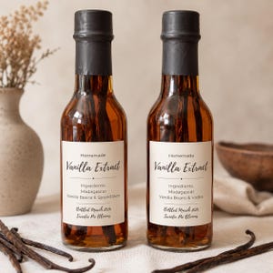 May include: Two clear glass bottles filled with amber liquid and whole vanilla beans. The labels read Homemade Vanilla Extract, Madagascar Vanilla Beans & Spiced Rum or Vodka, and Bottled March 2026 Sweetie Pie Blooms. Black wax seals the bottle caps.