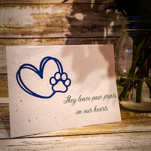 May include: A white card with a blue paw print inside a heart. The card has the text "They leave paw prints on our hearts".