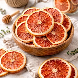 May include: A wooden bowl filled with dried orange slices, showcasing a vibrant red-orange color. The slices are arranged on a neutral-toned cloth, with additional slices scattered around. A glass jar filled with more slices is visible in the background.