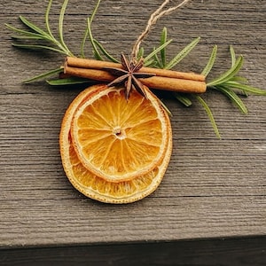Pode incluir: Um ornamento decorativo com duas fatias de laranja seca sobrepostas, cobertas por dois paus de canela castanhos e um anis estrelado castanho-escuro. Ramos de alecrim verde estendem-se por tr&aacute;s da canela, com um la&ccedil;o de corda de juta.