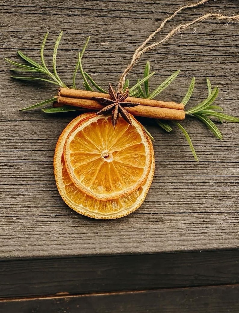 Pode incluir: Um ornamento decorativo com duas fatias de laranja seca sobrepostas, cobertas por dois paus de canela castanhos e um anis estrelado castanho-escuro. Ramos de alecrim verde estendem-se por tr&aacute;s da canela, com um la&ccedil;o de corda de juta.