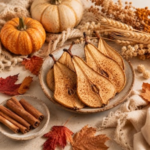 May include: A ceramic plate filled with several thin, dried pear slices dusted with cinnamon. Nearby, a smaller bowl holds cinnamon sticks, accompanied by two whole pumpkins, dried wheat stalks, and scattered autumn leaves on a textured fabric.