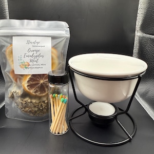 May include: A white ceramic simmer pot with a black metal stand, a clear bag of dried orange slices and herbs, and a small glass jar of matches with colorful tips. The bag is labeled "Stovetop Simmerpot Orange Eucalyptus Mint."