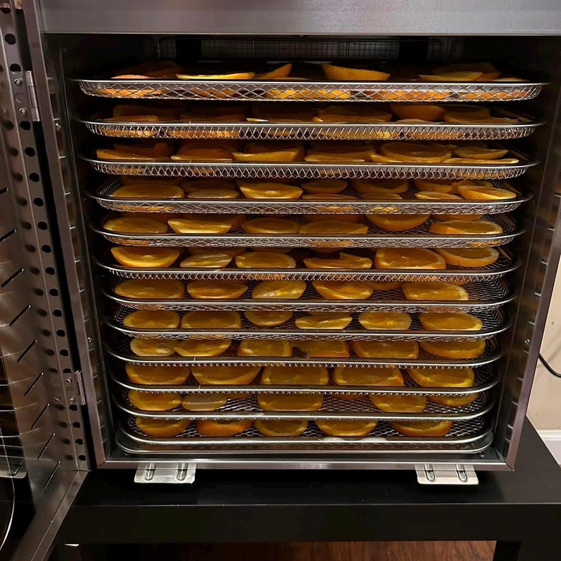 May include: A food dehydrator with multiple wire racks filled with sliced oranges. The open door reveals the drying process. The appliance is made of stainless steel. The image shows a method of preserving food, with the oranges arranged in rows.