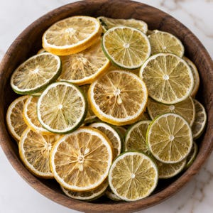 May include: A wooden bowl filled with dried lemon and lime slices. The slices vary in color from yellow to light brown and green, with visible textures and seeds. The bowl is made of dark wood and sits on a white marble surface.