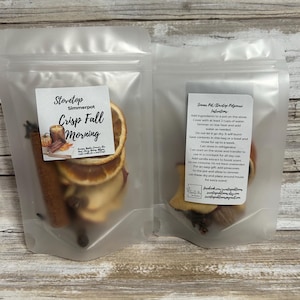 May include: Two clear plastic bags containing ingredients for a stovetop simmer pot. One bag features dried orange slices, cinnamon sticks, and other spices. The label reads "Stovetop Simmerpot Crisp Fall Morning."