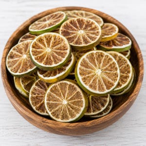May include: A wooden bowl filled with dried lime slices. The slices are circular, with a light brown centre and a green rind. The bowl is made of light brown wood and sits on a white wooden surface.