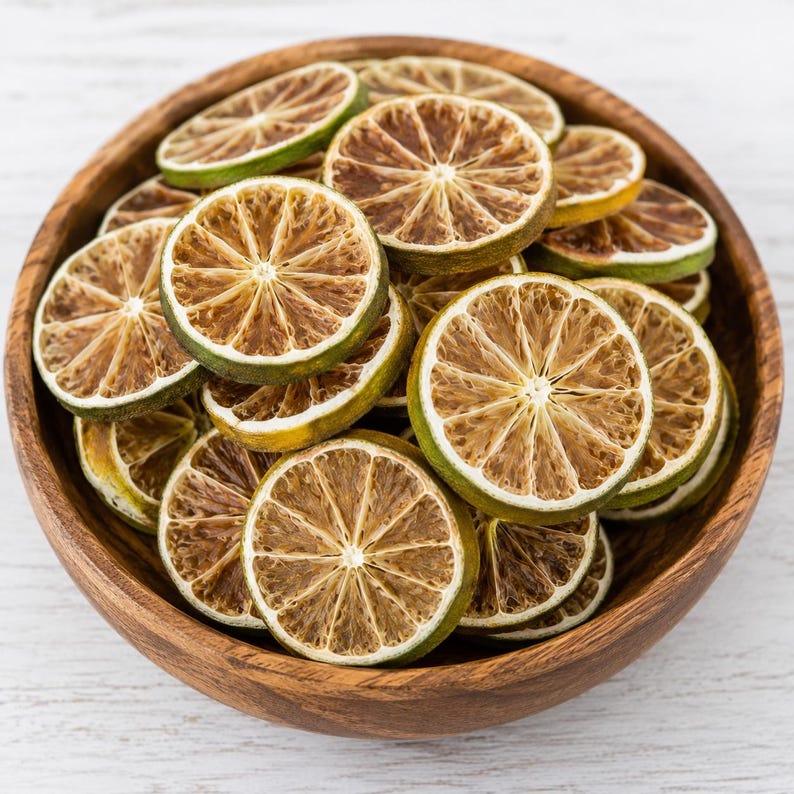 May include: A wooden bowl filled with dried lime slices. The slices are circular, with a light brown centre and a green rind. The bowl is made of light brown wood and sits on a white wooden surface.
