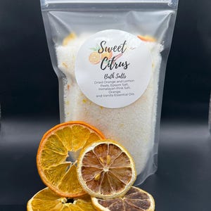 May include: A clear bag of Sweet Citrus bath salts with a round label. The label lists ingredients like dried orange and lemon peels. Several dried orange and lemon slices are in front of the bag. The background is black.