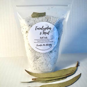 May include: A clear plastic bag filled with white bath salts, eucalyptus leaves, and a white label that reads "Eucalyptus & Mint Bath Salts Dried Eucalyptus & Mint Leaf, Epsom and Sea Salt Mint & Eucalyptus Essential Oils Sweetie Pie Blooms sweetiepieblooms.etsy.com".