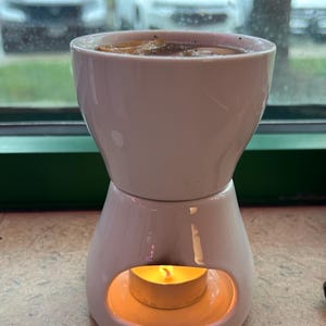May include: A white ceramic oil warmer with a candle burning inside. The warmer has a two-part design, with a cup-shaped top and a base with an opening for the candle. The warmer is sitting on a textured surface.