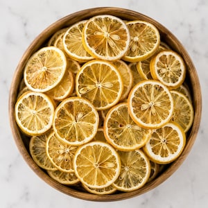 May include: A wooden bowl filled with dried lemon slices. The slices are yellow and tan, with visible seeds and a circular shape. The bowl is made of light brown wood and sits on a white surface. The lemons are arranged in a circular pattern.