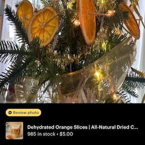 Pode incluir: Uma guirlanda de fatias de laranja desidratadas em um barbante, exibida como decora&ccedil;&atilde;o em uma &aacute;rvore de Natal verde com fita dourada e luzes brancas quentes. As fatias de laranja circulares e transl&uacute;cidas est&atilde;o dispostas entre os ramos.