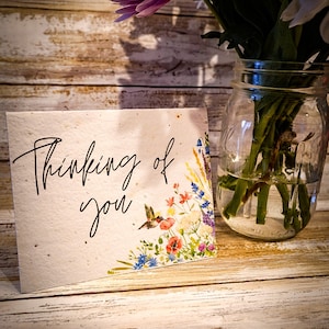 May include: A card with the text "Thinking of you" in black script on a white background. The card has a floral design with a hummingbird and colorful flowers.