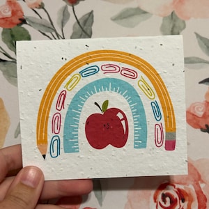 May include: A rectangular card with a rainbow design featuring a red apple, paper clips, and a pencil. The rainbow arches over a ruler design. The card is white with small flecks, and the apple has a smiling face.