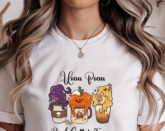 May include: White t-shirt with a Halloween-themed graphic. The design features three cartoon coffee drinks with witch-like hair and the text "Hocus Pocus Need Coffee to Focus". The shirt is worn by a person.