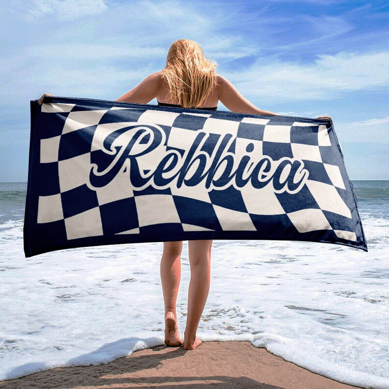May include: A navy blue and white checkered beach towel with the name "Rebbica" in a cursive font. The towel is held up by a person on a sandy beach with the ocean in the background.