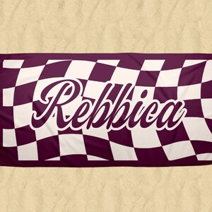 May include: A rectangular beach towel with a checkered pattern in burgundy and white. The name "Rebbica" is displayed in a white, cursive font across the center of the towel. The towel is laid out on a sandy surface.