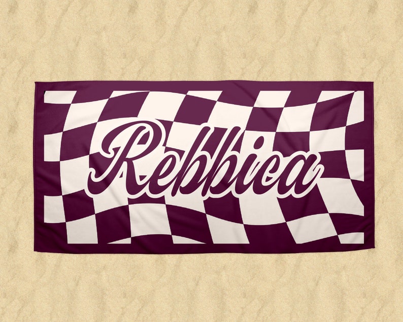 May include: A rectangular beach towel with a checkered pattern in burgundy and white. The name "Rebbica" is displayed in a white, cursive font across the center of the towel. The towel is laid out on a sandy surface.