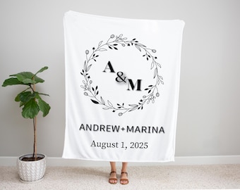 May include: White blanket with a black floral wreath design, the initials "A & M", and the names "ANDREW + MARINA" with the date "August 1, 2025". The blanket is held up in front of a light gray wall.