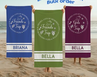 May include: Three beach towels in blue, green, and burgundy, each held by a person on a sandy beach. The towels feature a circular design with the words "Friends Trip" or "Girls Trip" and the year 2026. Each towel has a white stripe with a name.