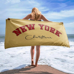 May include: A yellow beach towel with the text "NEW YORK" in red and the name "Christina" in black cursive. The towel is being held by a person on a beach.