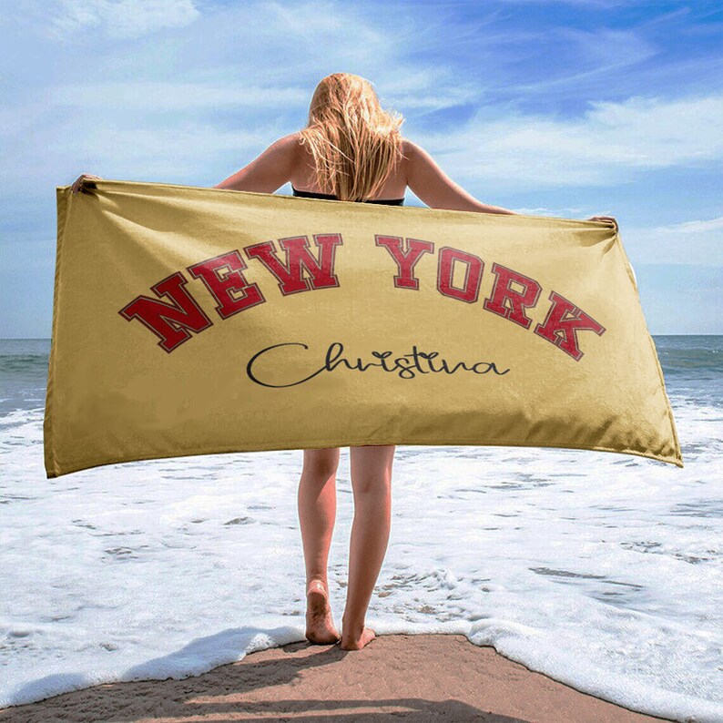 May include: A yellow beach towel with the text "NEW YORK" in red and the name "Christina" in black cursive. The towel is being held by a person on a beach.