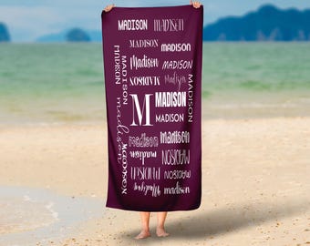 May include: A dark purple beach towel with the name "Madison" printed in white in various fonts and sizes. The towel is held up against a beach backdrop with sand, water, and a distant shoreline.