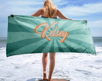 May include: A teal beach towel with a retro sunburst design and the name "Kelsey" in orange script. The towel is held up by a person on a sandy beach with the ocean in the background.