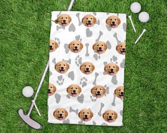 May include: White golf towel featuring a pattern of smiling golden retriever puppy faces, gray paw prints, and bones. The towel is on green grass with golf balls, tees, and a putter. A fun accessory for dog lovers who enjoy golf.